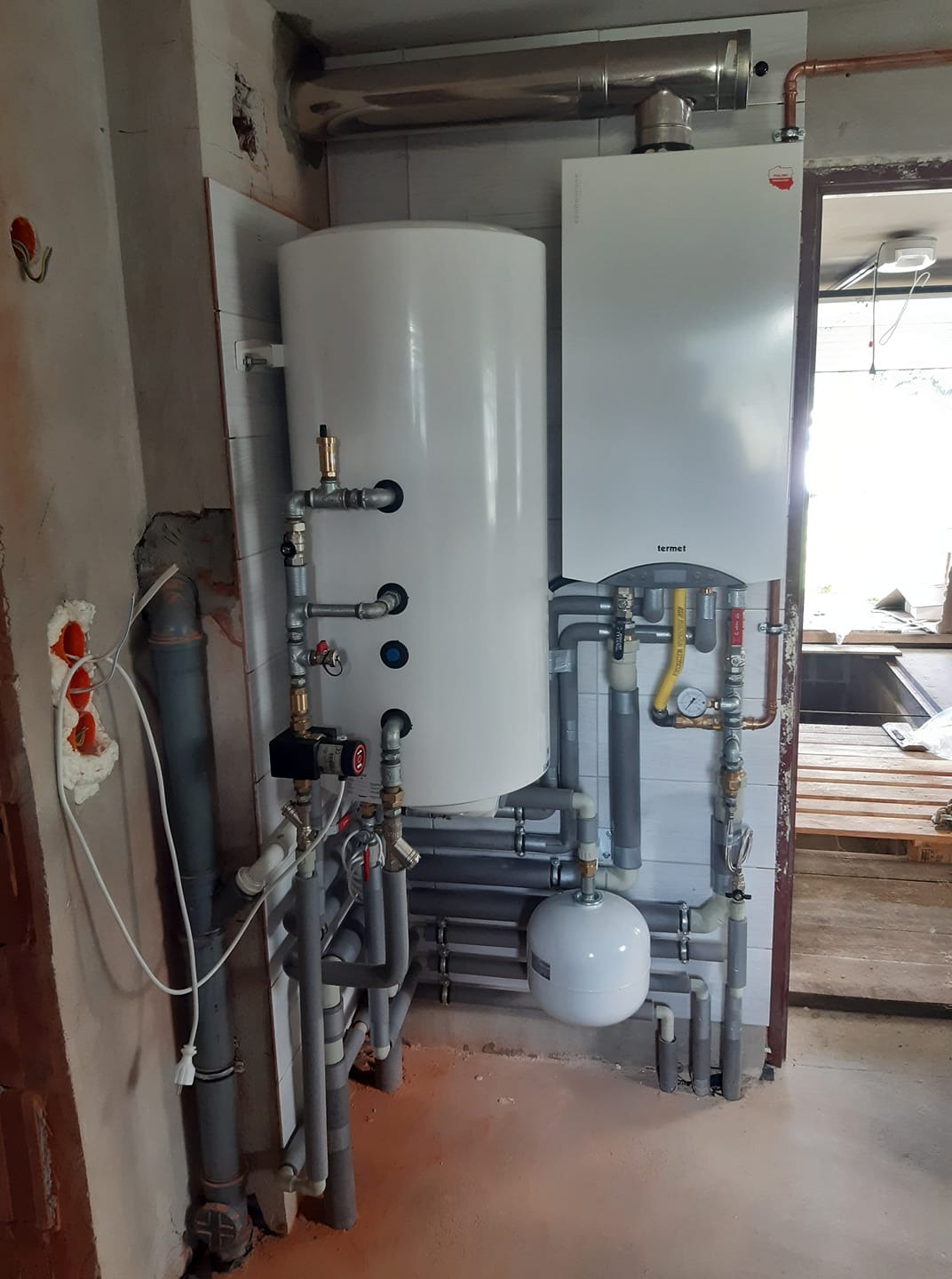 Biały zasobnik ciepłej wody i piec gazowy marki Termet, połączone licznymi rurami z zaworami, zamontowane przy ścianie z częściowo położonymi białymi płytkami, widoczne ślady prac wykończeniowych.
