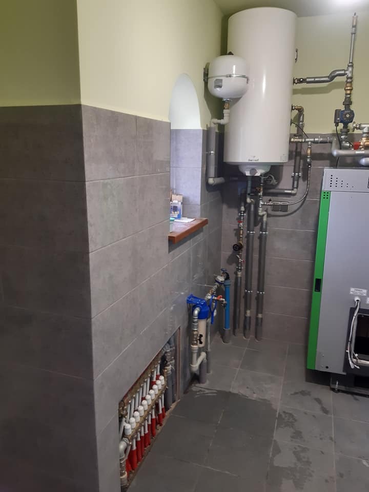 Kotłownia z piecem, zasobnikiem ciepłej wody, rozdzielaczem ogrzewania podłogowego i filtrem wody, szare płytki na ścianach i podłodze, widoczny fragment instalacji grzewczej.