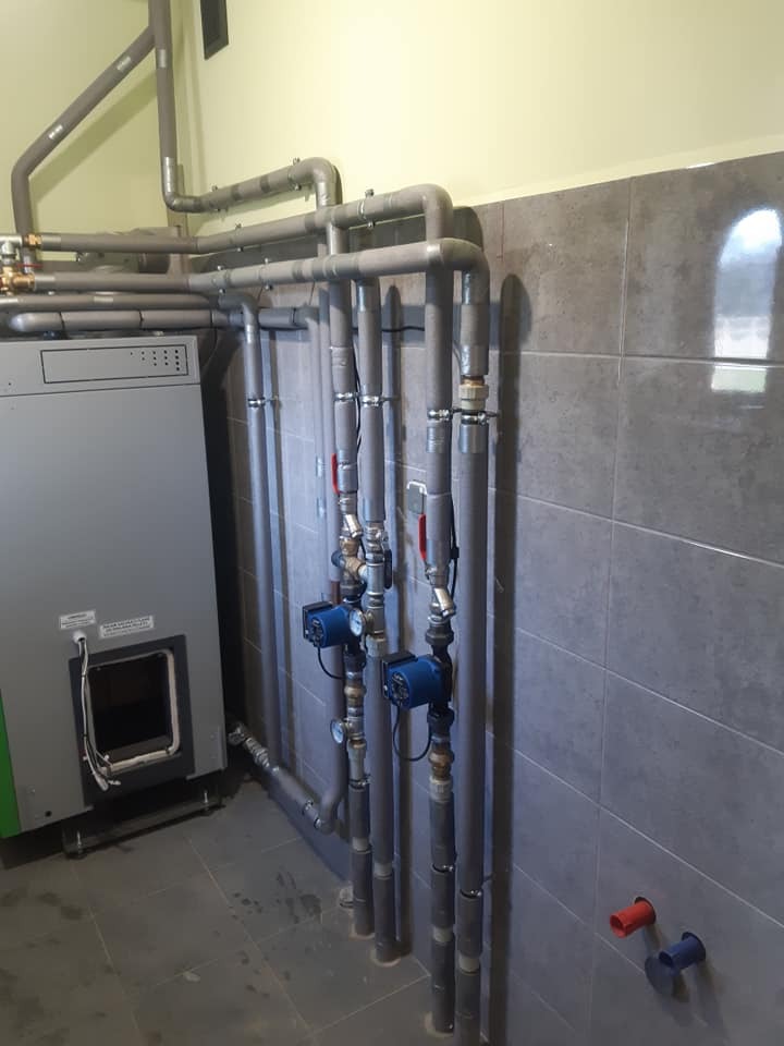 Nowoczesna kotłownia z piecem na paliwo stałe, rurami w izolacji i pompami obiegowymi na tle szarych płytek ceramicznych.