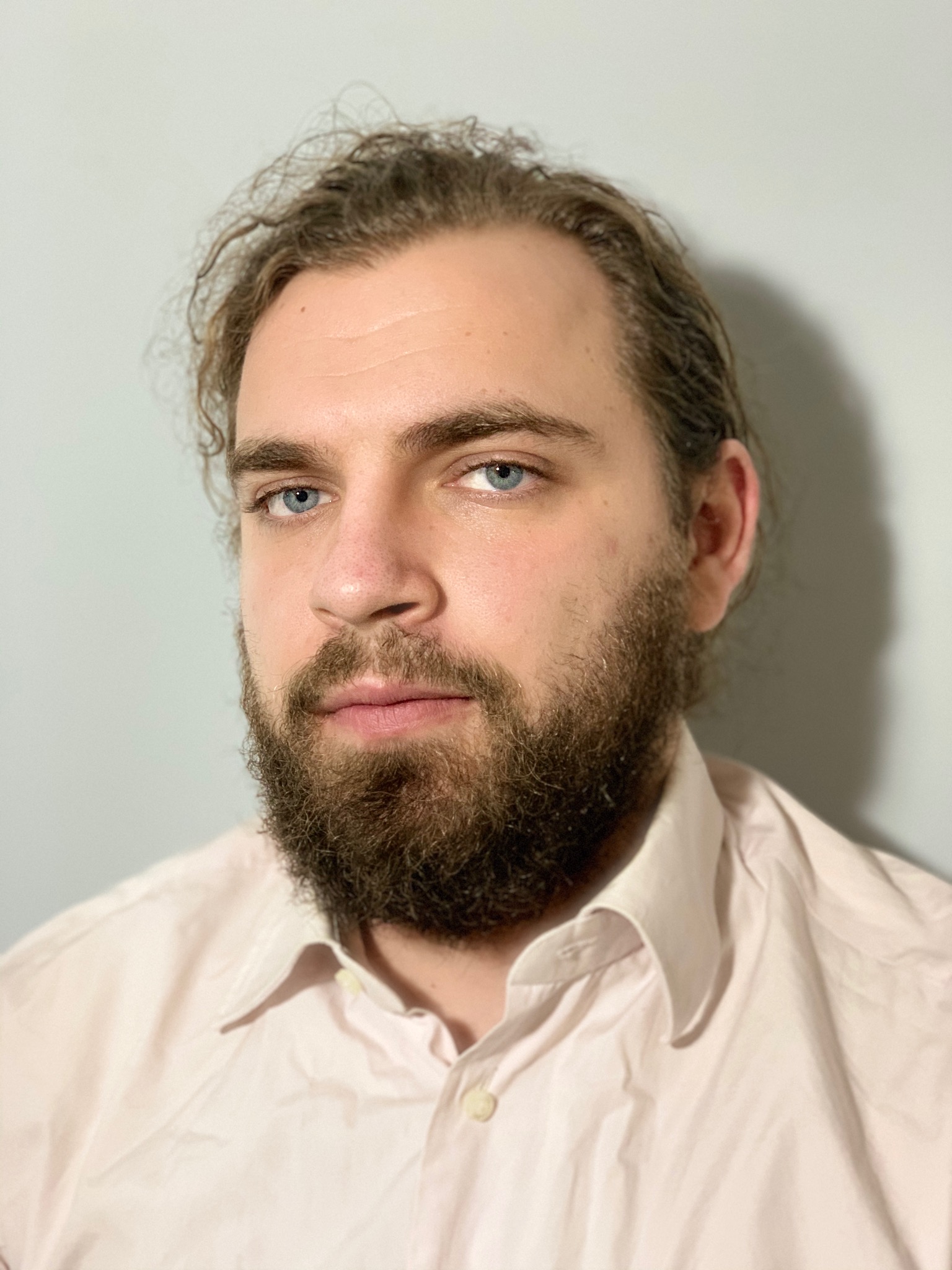 Portret brodatego mężczyzny z niebieskimi oczami w jasnej koszuli na tle szarej ściany.