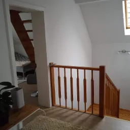 Jasne wnętrze domu z drewnianymi schodami i balustradą, widok na fragment pokoju z łóżkiem przez otwarte drzwi, na ścianie dekoracyjna grafika z napisem.