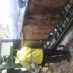 Pracownik w żółtej kamizelce obsługuje traka taśmowego, tnąc masywny drewniany blok na deski. Widoczna surowa faktura drewna i elementy konstrukcyjne maszyny.