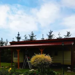 Domek letniskowy z czerwonym dachem i drewnianą konstrukcją, malowany przez mężczyznę stojącego na drabinie, otoczony zielonym ogrodem z kwitnącymi kwiatami.