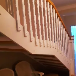 Białe schody z drewnianą balustradą, widok od dołu, pod schodami widoczne krzesła w folii ochronnej i karton.