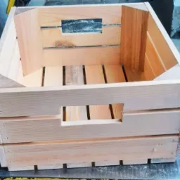 Surowa, jasna, drewniana skrzynka z uchwytami, wykonana z desek połączonych gwoździami, ustawiona na warsztatowym stole.