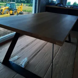 Stół z blatem z litego drewna o ciemnym wybarwieniu, wsparty na metalowej, geometrycznej podstawie, widok z boku, w tle okno z widokiem na plac budowy z koparką.