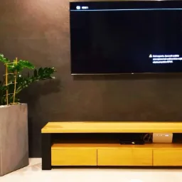 Nowoczesna komoda RTV z jasnego drewna z trzema szufladami i czarnymi nogami, umieszczona pod wiszącym telewizorem na tle ściany w odcieniach szarości, obok duża donica z zieloną rośliną.