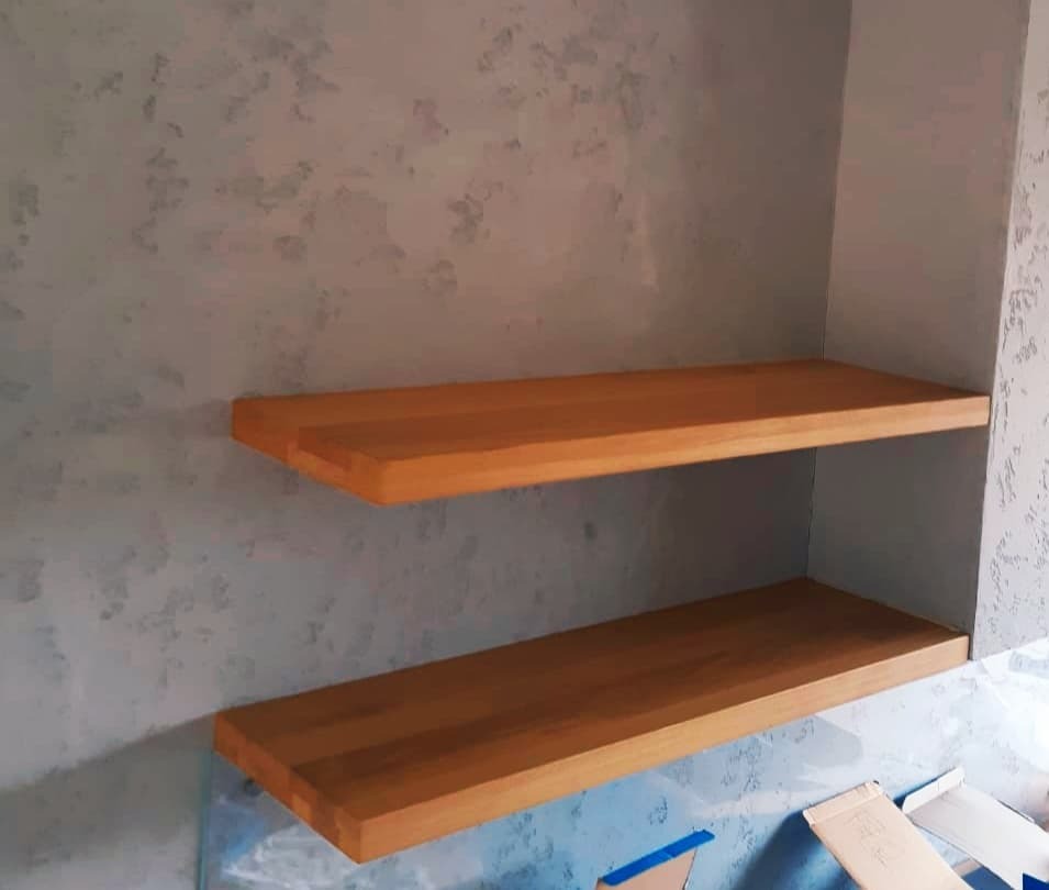 Dwie proste, drewniane półki z jasnego drewna zamontowane na szarej ścianie z dekoracyjnym wzorem. Widoczne ślady prac wykończeniowych.