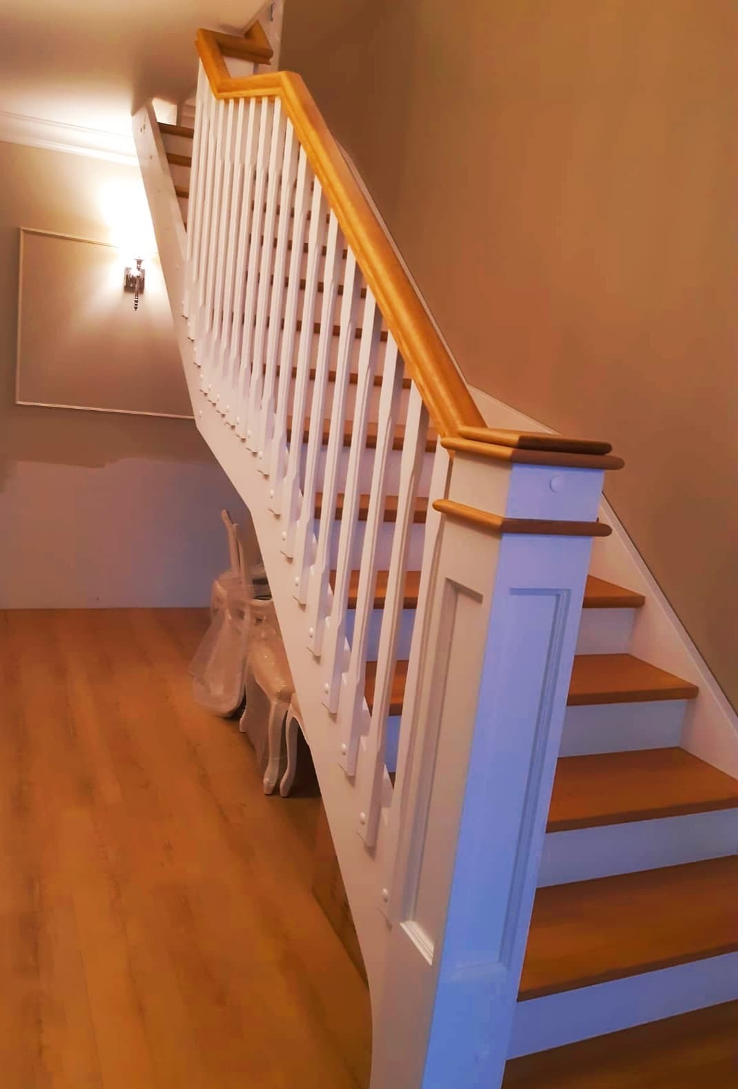 Drewniane schody z białymi balustradami i złoconym poręczą, widoczne krzesła zabezpieczone folią ochronną, oświetlenie ścienne w tle.