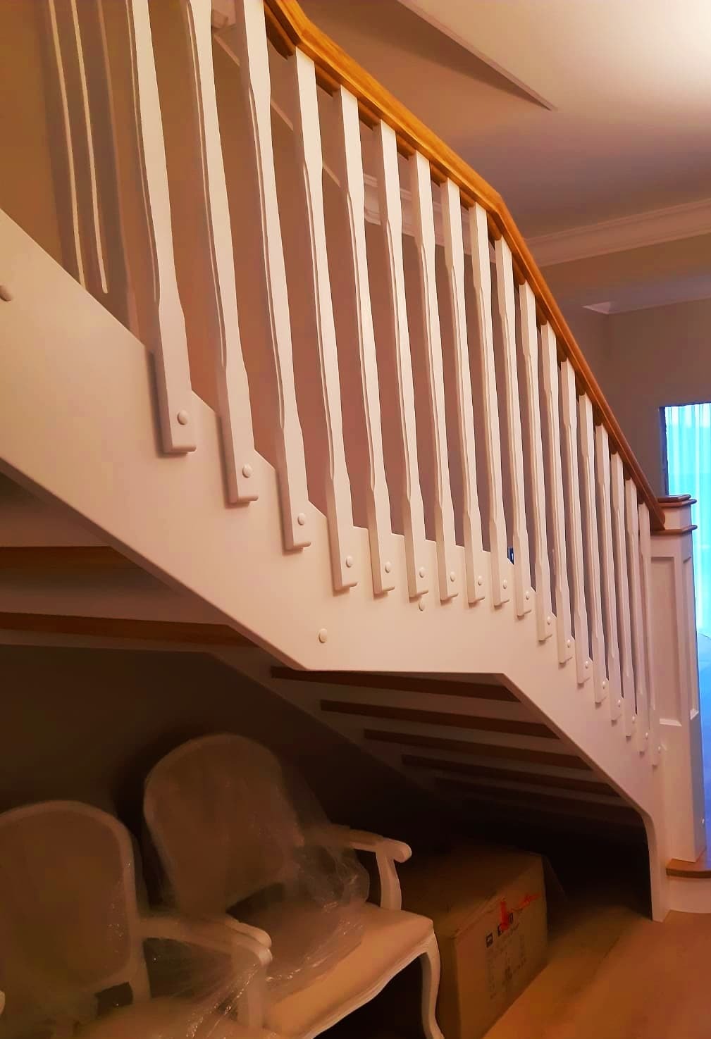 Białe schody z drewnianą balustradą, widok od dołu, pod schodami widoczne krzesła w folii ochronnej i karton.