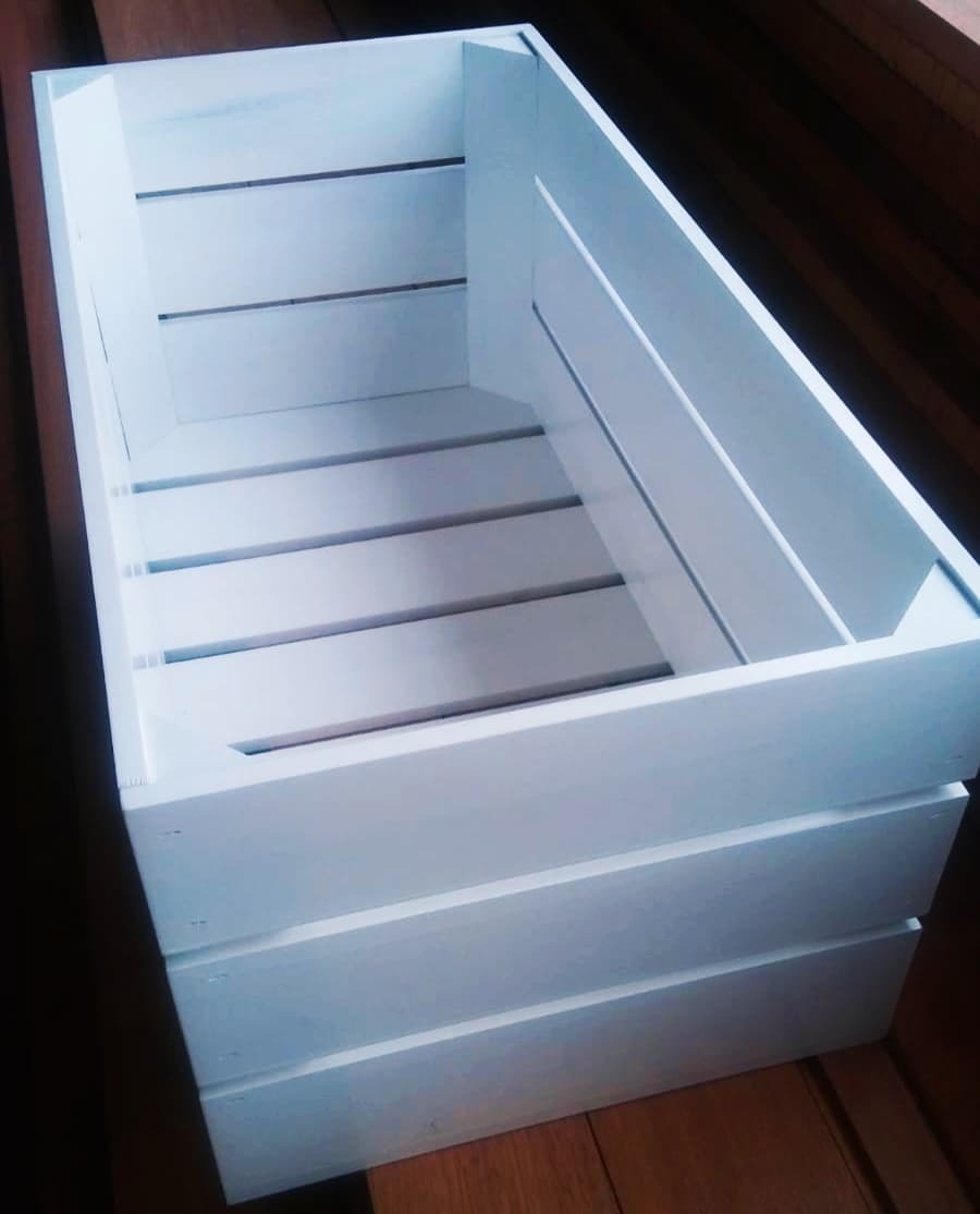 Biała, drewniana skrzynka z listewek, widoczna z góry, na drewnianej podłodze.