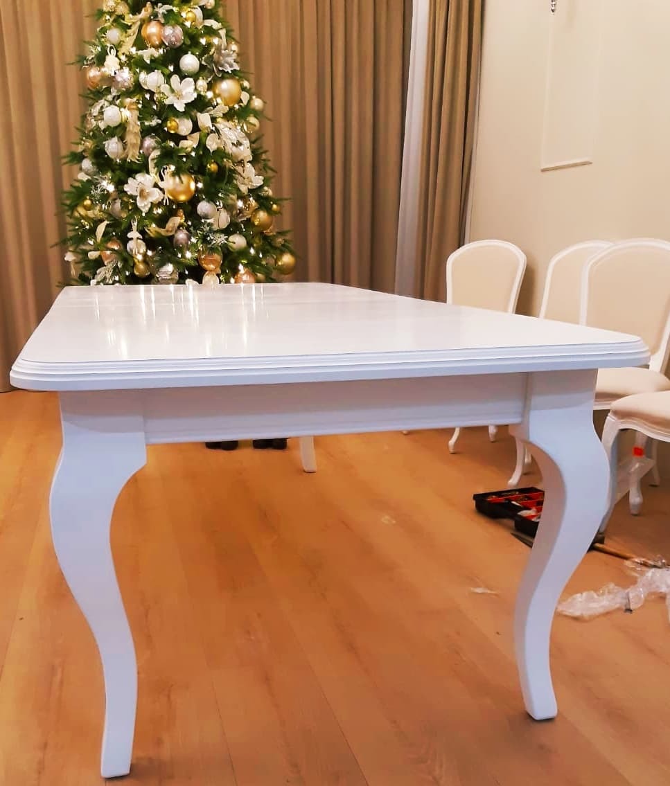 Biały, błyszczący stół na giętych nogach w stylu ludwikowskim, częściowo zmontowany, z choinką w tle i narzędziami na podłodze.