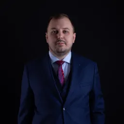 Portret mężczyzny w granatowym garniturze z bordowym krawatem na czarnym tle.