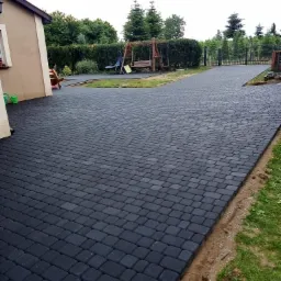 Nowoczesna, ciemnoszara nawierzchnia z kostki brukowej prowadząca od domu do bramy w ogrodzeniu, z placem zabaw i huśtawką w tle.