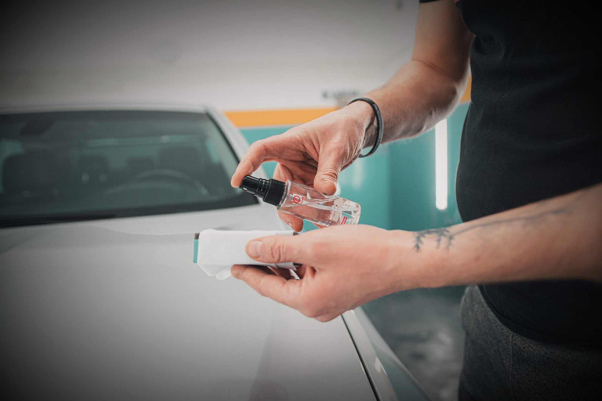 Nakładanie płynu z atomizera na aplikator z mikrofibry, przygotowanie do polerowania lakieru samochodu, widoczne dłonie mężczyzny z tatuażem.