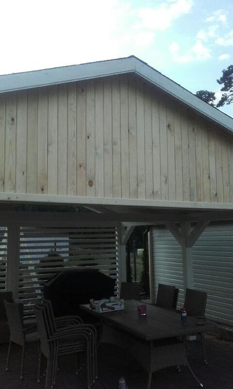 Zadaszenie tarasu z jasnego drewna z widocznym stołem i krzesłami w przestrzeni pod nim, w tle widoczny budynek z szarej blachy falistej.