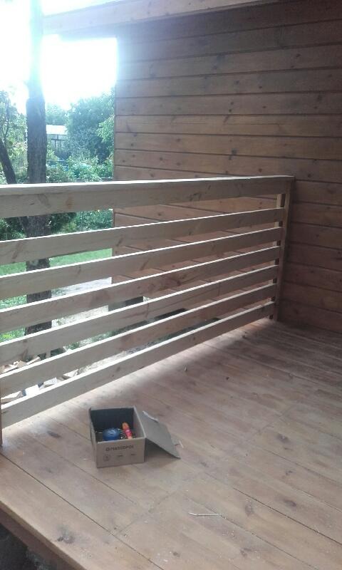 Wnętrze drewnianego tarasu z niepomalowaną balustradą, widok na zieleń przez szczeliny, kartonowe pudełko z narzędziami w rogu.