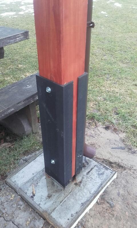 Drewniany słup z czarnymi osłonami mocowany na betonowej podstawie, obok rynna spustowa, widoczny fragment ławki i trawnik.