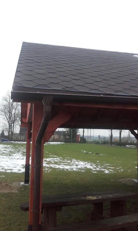 Zadaszenie altany pokryte ciemną dachówką bitumiczną, widoczne rynny i drewniana konstrukcja, w tle trawnik z resztkami śniegu.