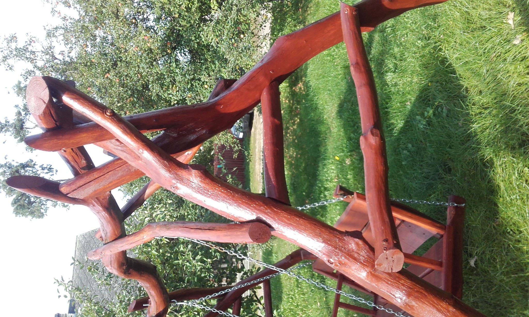 Drewniana huśtawka ogrodowa wykonana z gałęzi, zawieszona na łańcuchach, z widocznym trawnikiem i drzewami w tle, polakierowana na ciemny brąz.