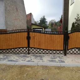 Dwuskrzydłowa brama wjazdowa z drewnianym wypełnieniem i metalową ramą o dekoracyjnym wzorze rombów, osadzona na świeżo ułożonej kostce brukowej, z widocznymi budynkami mieszkalnymi w tle.