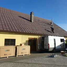 Montaż konstrukcji pod panele słoneczne na brązowym dachu domu z żółtą elewacją, widoczne dwie duże paczki z panelami Q CELLS, biały van z otwartymi drzwiami i drabina oparta o dach.