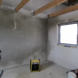 Surowe wnętrze w trakcie prac wykończeniowych: świeżo otynkowane ściany, drewniane belki stropowe, okno PCV z nieusuniętą folią ochronną, żółty grzejnik elektryczny i lampa budowlana na statywie.