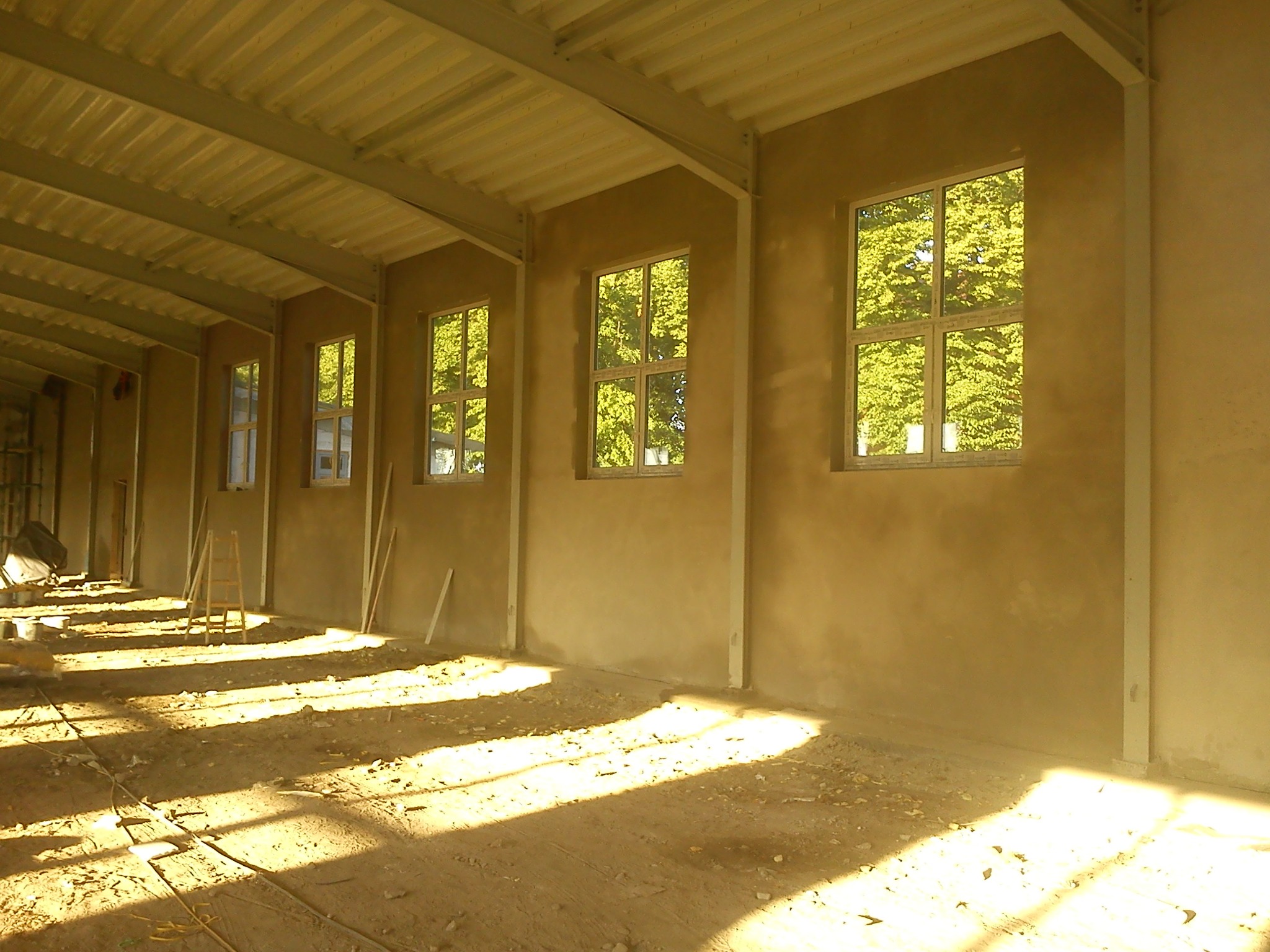 Wnętrze budynku w trakcie tynkowania maszynowego, widoczne surowe ściany z oknami z widokiem na drzewa, metalowa konstrukcja dachu oraz podłoga z resztkami zaprawy i narzędziami, oświetlone silnym...