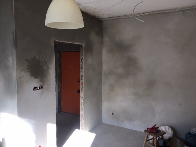 Świeżo otynkowane ściany wewnątrz pomieszczenia, widoczne ślady po nałożonym tynku, otwór drzwiowy bez ościeżnicy, na podłodze narzędzia i krzesło.