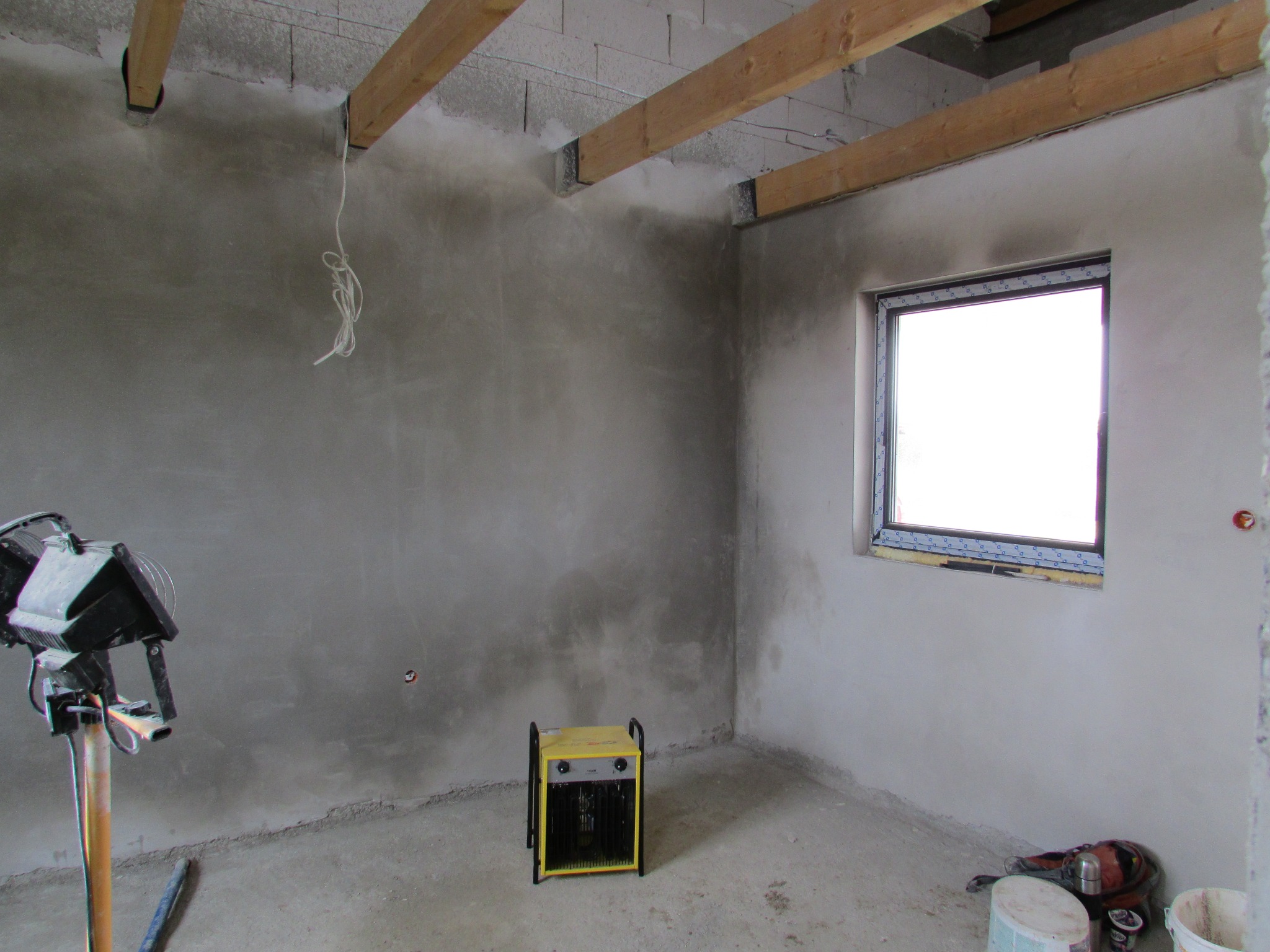 Surowe wnętrze w trakcie prac wykończeniowych: świeżo otynkowane ściany, drewniane belki stropowe, okno PCV z nieusuniętą folią ochronną, żółty grzejnik elektryczny i lampa budowlana na statywie.