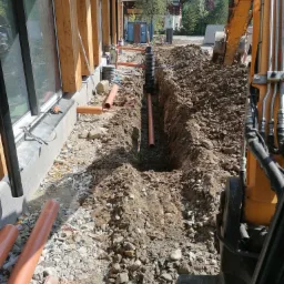 Wykopany rów wzdłuż budynku z widocznymi rurami kanalizacyjnymi w ziemi i pomarańczowymi rurami na powierzchni, z żółtą koparką w tle i elementami krajobrazu w oddali.