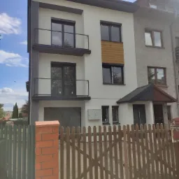 Nowoczesny dom z białą elewacją, szarymi akcentami i drewnianymi panelami, widoczny zza drewnianego płotu