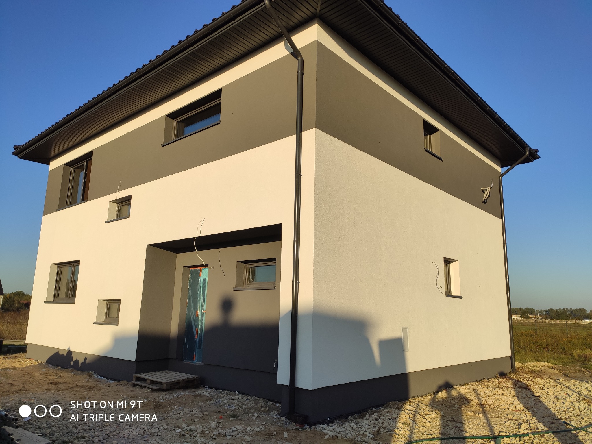 Dwukondygnacyjny dom z elewacją w odcieniach szarości i bieli, widoczne okna i rynna, teren wokół domu częściowo zagospodarowany, drzwi wejściowe zabezpieczone folią.