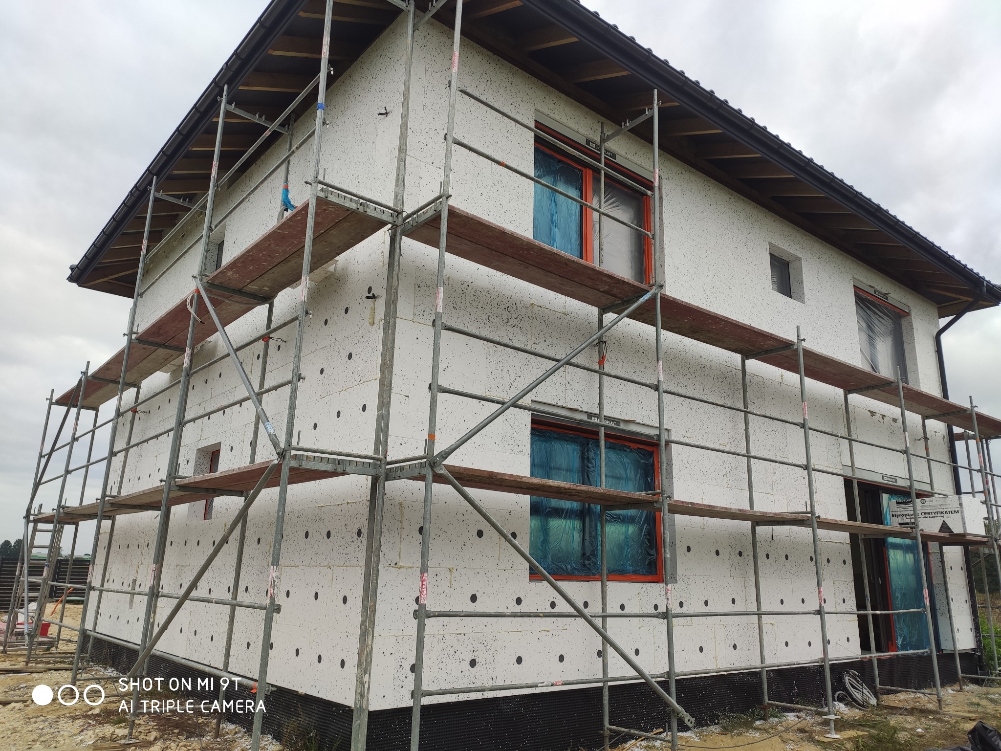 Dom w trakcie ocieplania elewacji styropianem, widoczne rusztowania i okna zabezpieczone folią, czarne kołki mocujące izolację.