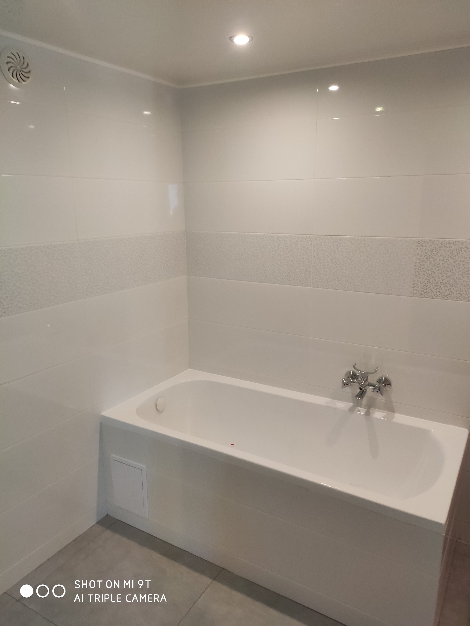 Nowoczesna, biała wanna obudowana panelami, z chromowaną baterią, na tle białych płytek z dekoracyjnym pasem, widoczny wentylator w rogu, oświetlenie sufitowe.