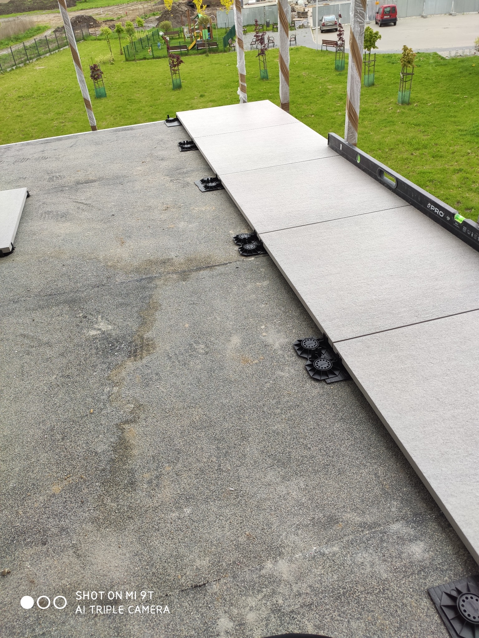 Układanie szarych płyt tarasowych na regulowanych wspornikach z użyciem poziomicy, widok z góry na plac zabaw w tle.