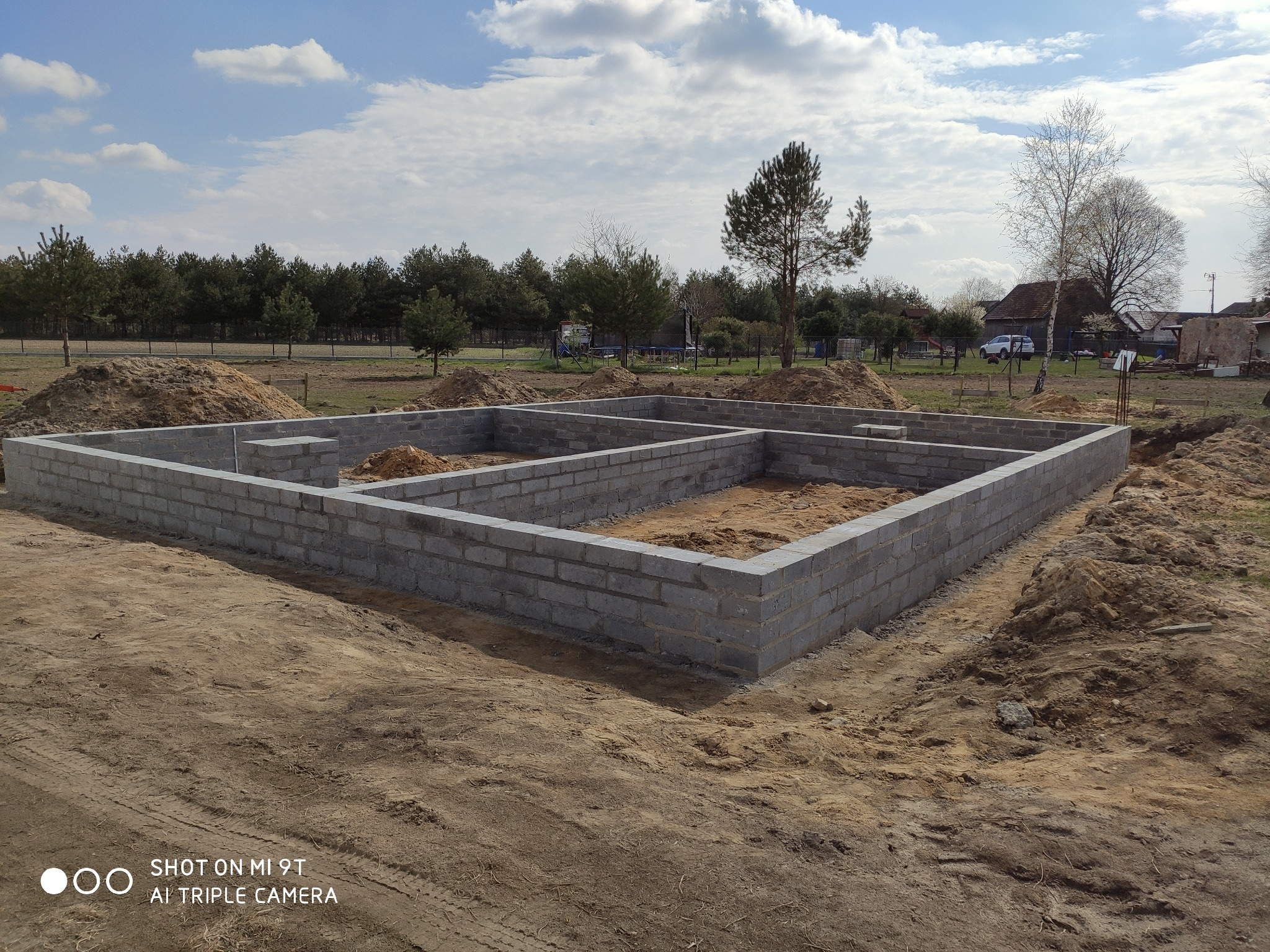 Wzniesione fundamenty z bloczków betonowych na piaszczystym terenie budowy, widoczne pryzmy piasku i drzewa w tle pod błękitnym niebem.