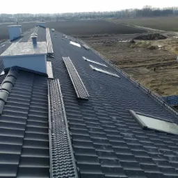 Widok z góry na nowy dach z ciemnej dachówki ceramicznej z zamontowanymi oknami dachowymi, kominami i metalowymi pomostami komunikacyjnymi na tle krajobrazu z polami i budynkami w oddali.