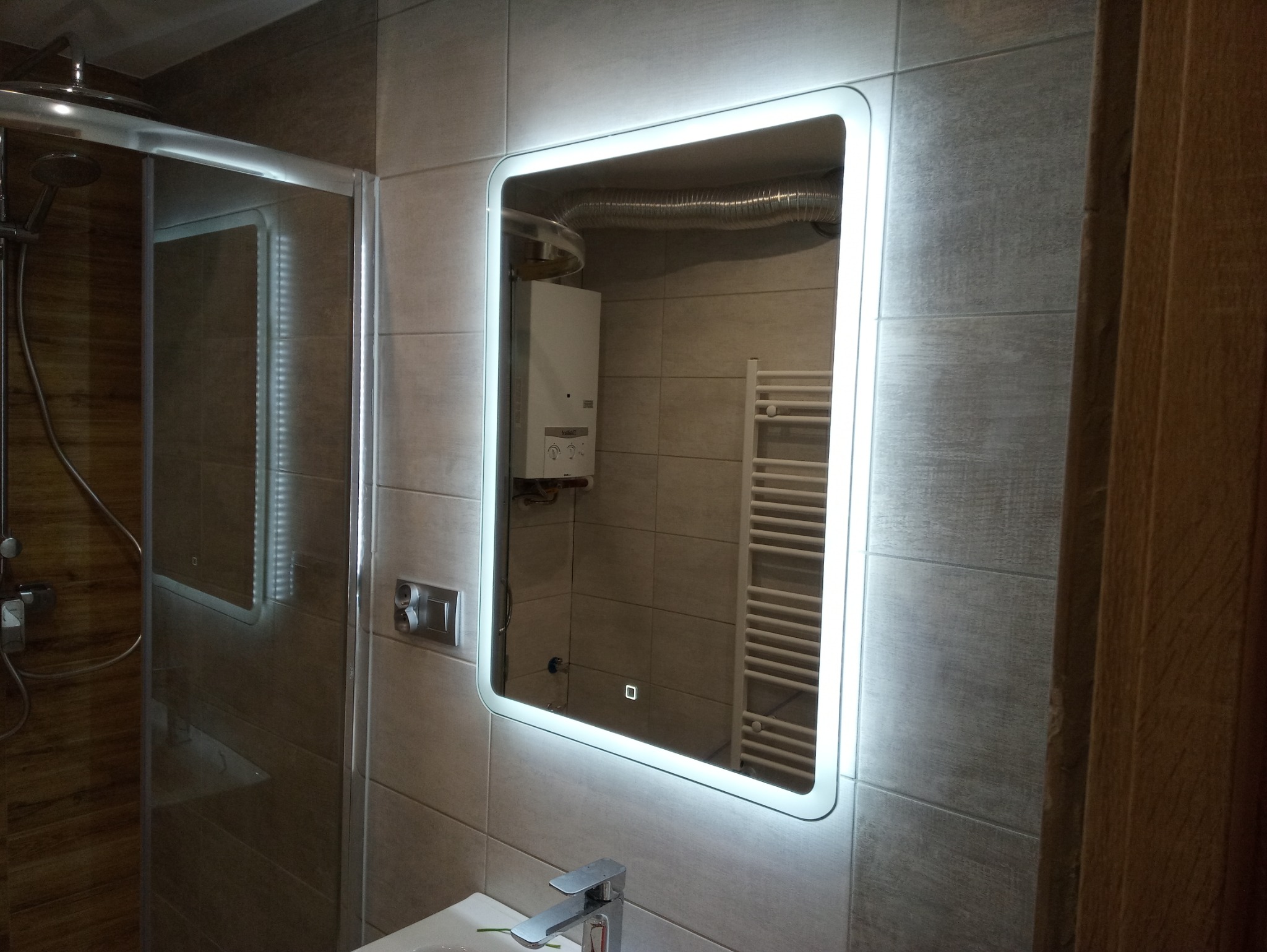 Wykończona łazienka z podświetlanym lustrem, widoczna kabina prysznicowa, grzejnik drabinkowy oraz piecyk gazowy w odbiciu lustra.