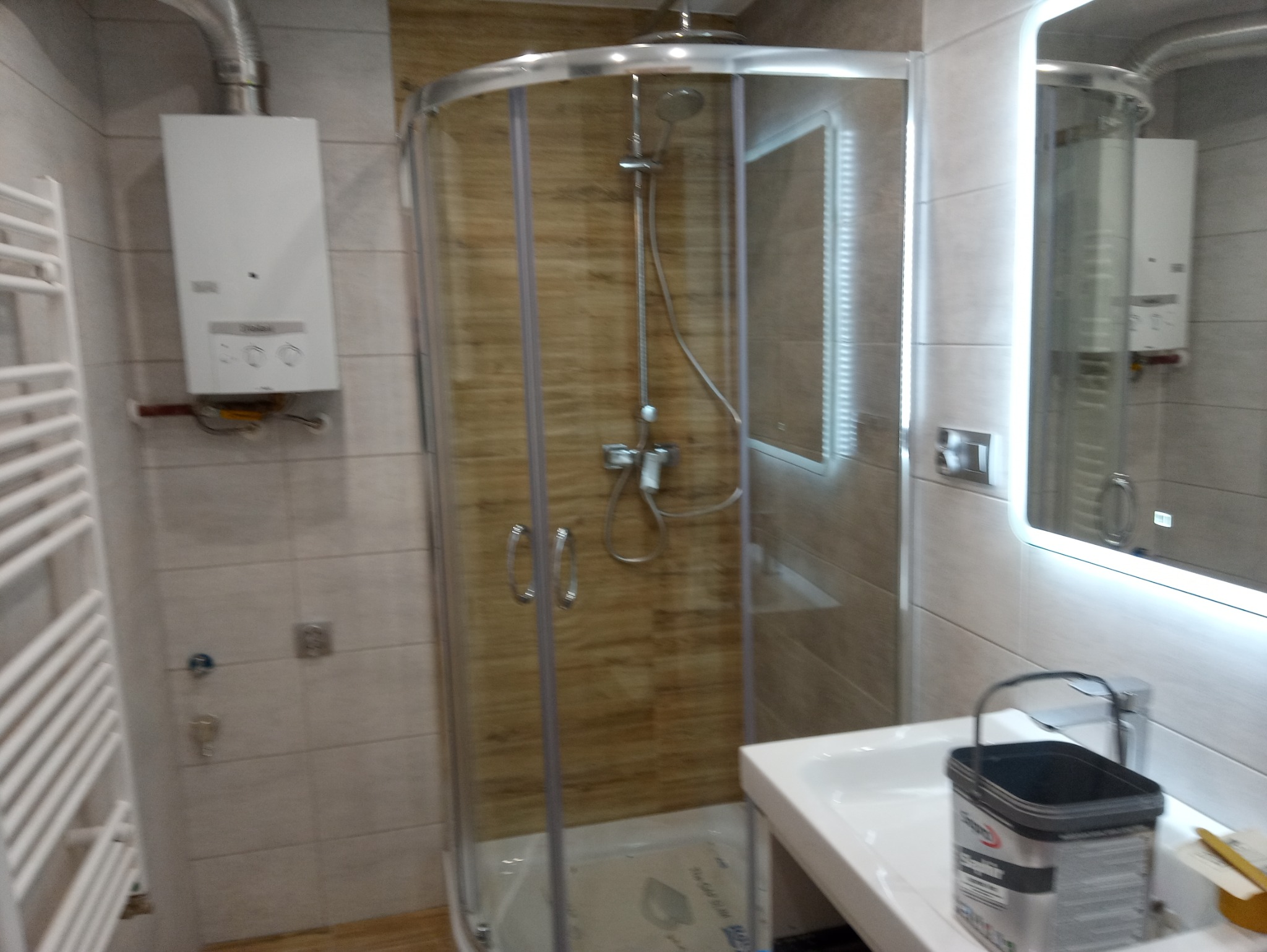 Wykończona łazienka z kabiną prysznicową z drewnopodobnymi płytkami, podgrzewaczem wody na ścianie, grzejnikiem drabinkowym i umywalką z wiadrem farby na blacie.