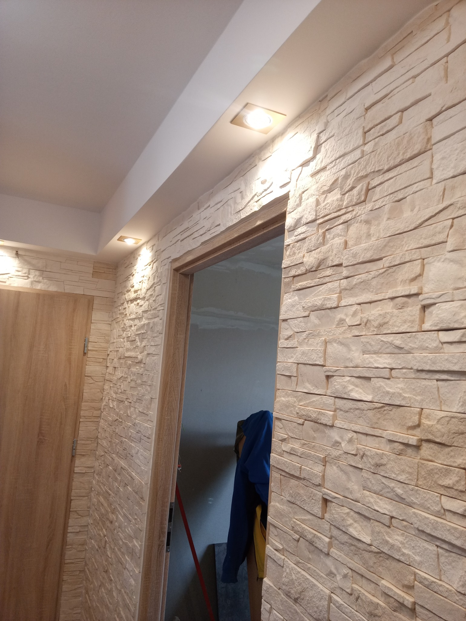 Korytarz z dekoracyjną ścianą wyłożoną jasnymi płytkami imitującymi kamień, oświetlony punktowymi lampami w suficie podwieszanym, widoczne drzwi z ościeżnicą również wykończoną okładziną kamienną.