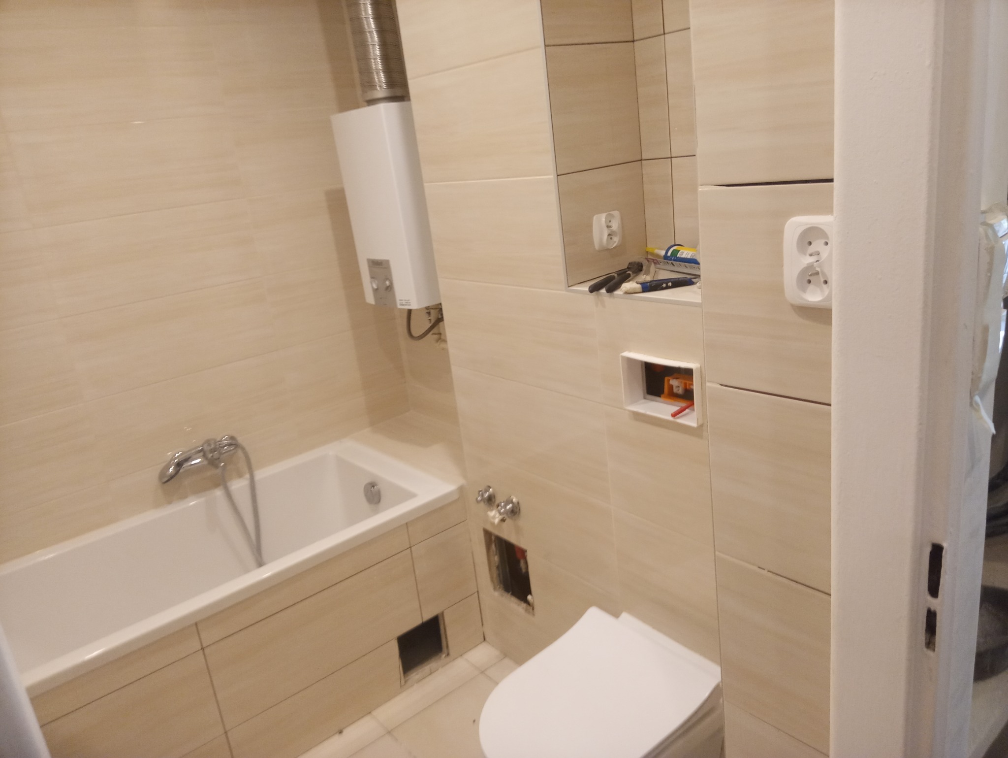 Łazienka w trakcie remontu: beżowe płytki, wanna, toaleta, bojler i niedokończone instalacje elektryczne oraz hydrauliczne.