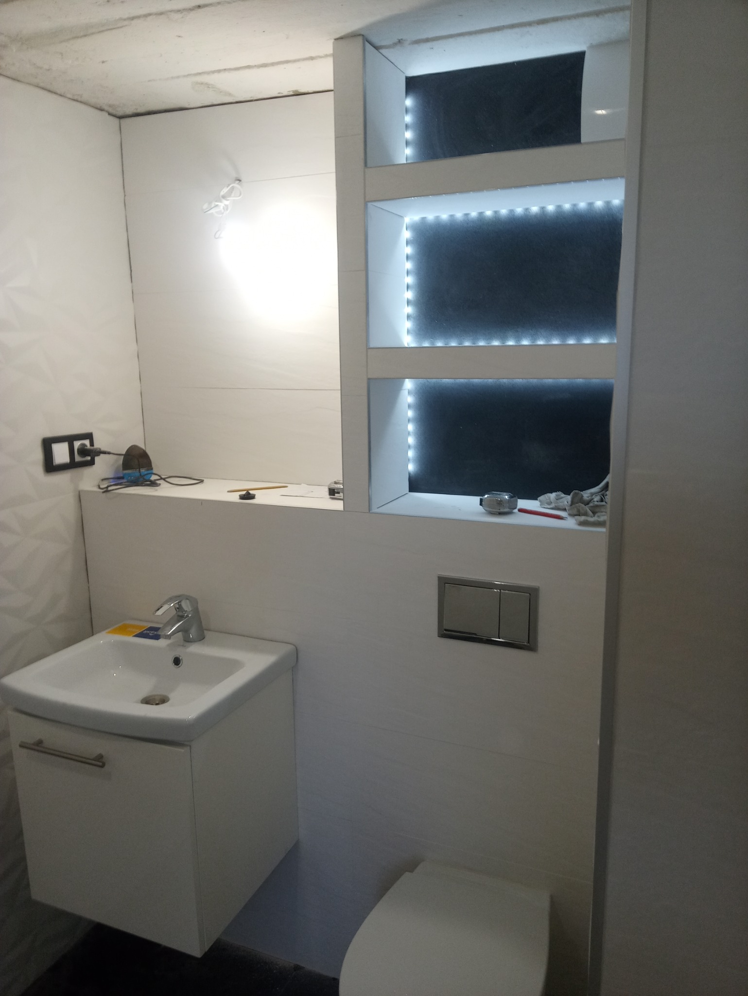 Biała łazienka po remoncie z umywalką, szafką pod umywalką, toaletą, lustrem z półkami podświetlanymi LED i żelazkiem na półce.