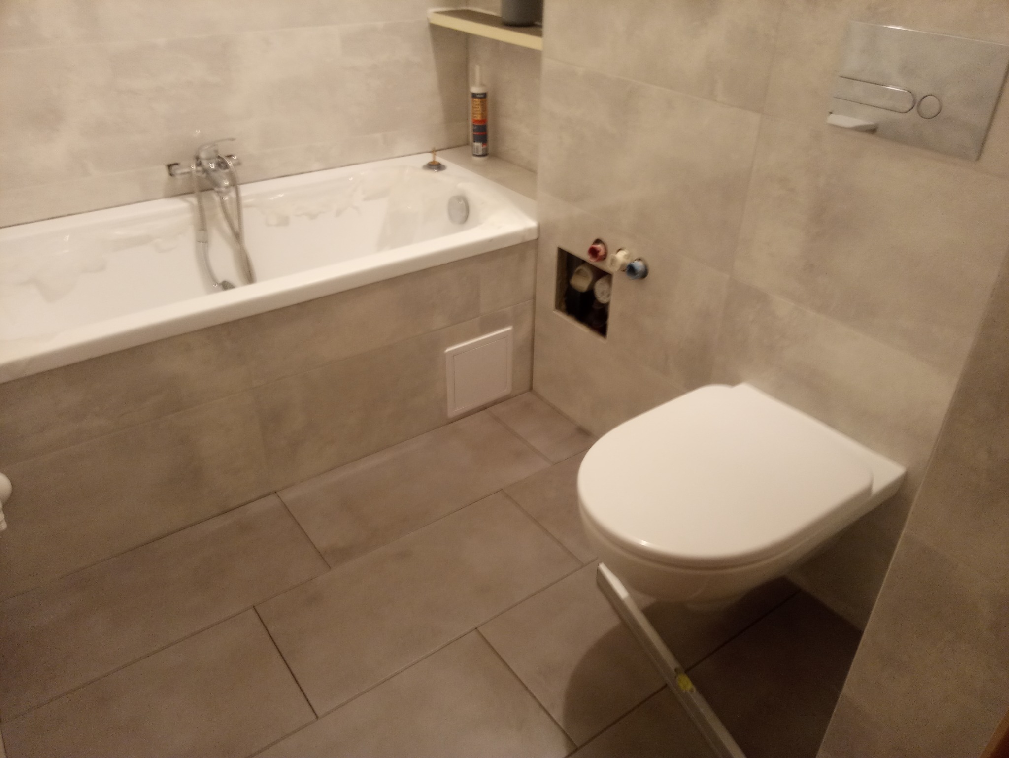 Wykończona łazienka z wanną obudowaną płytkami, wiszącą toaletą i szarą glazurą na ścianach i podłodze. Widoczne przyłącza wodne i biały pojemnik z silikonem na półce.