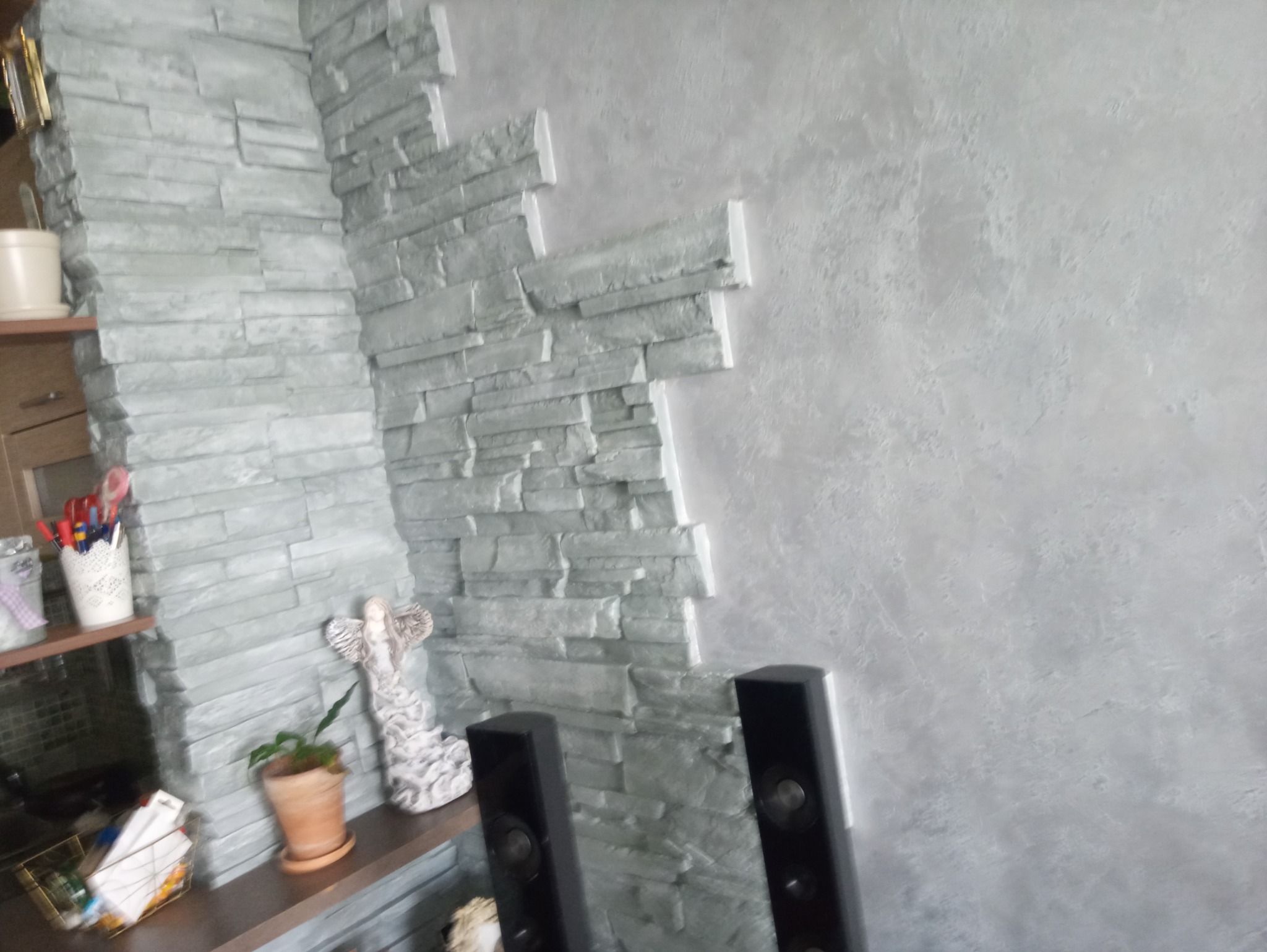 Fragment ściany z dekoracyjnym wykończeniem: imitacja kamienia w odcieniach szarości łączy się z gładką, jasnoszarą powierzchnią. Na półce widoczna figurka anioła i doniczka z rośliną.