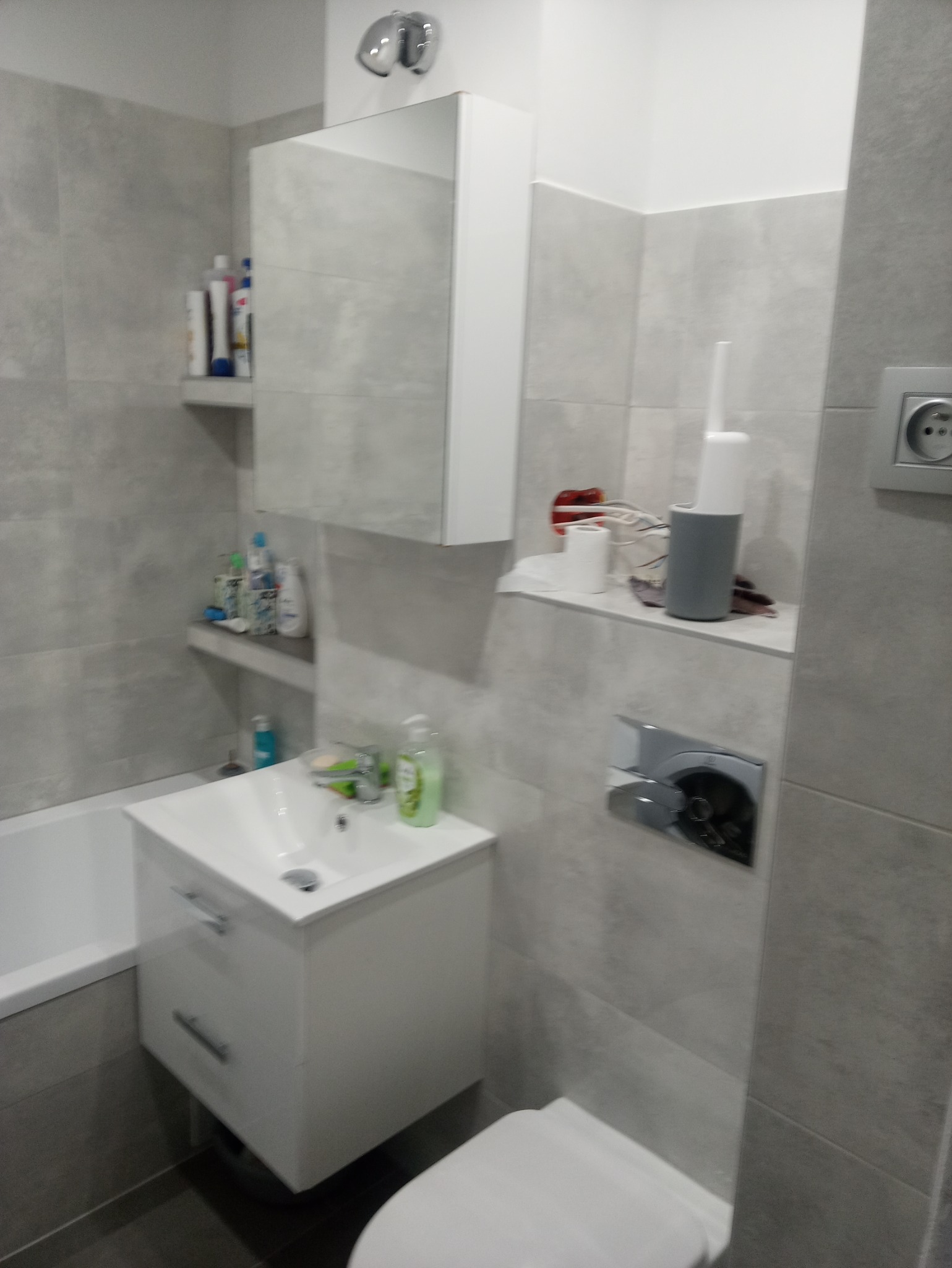 Szara łazienka po remoncie z białą szafką pod umywalką, baterią, toaletą i płytkami imitującymi beton, z półką na akcesoria łazienkowe.