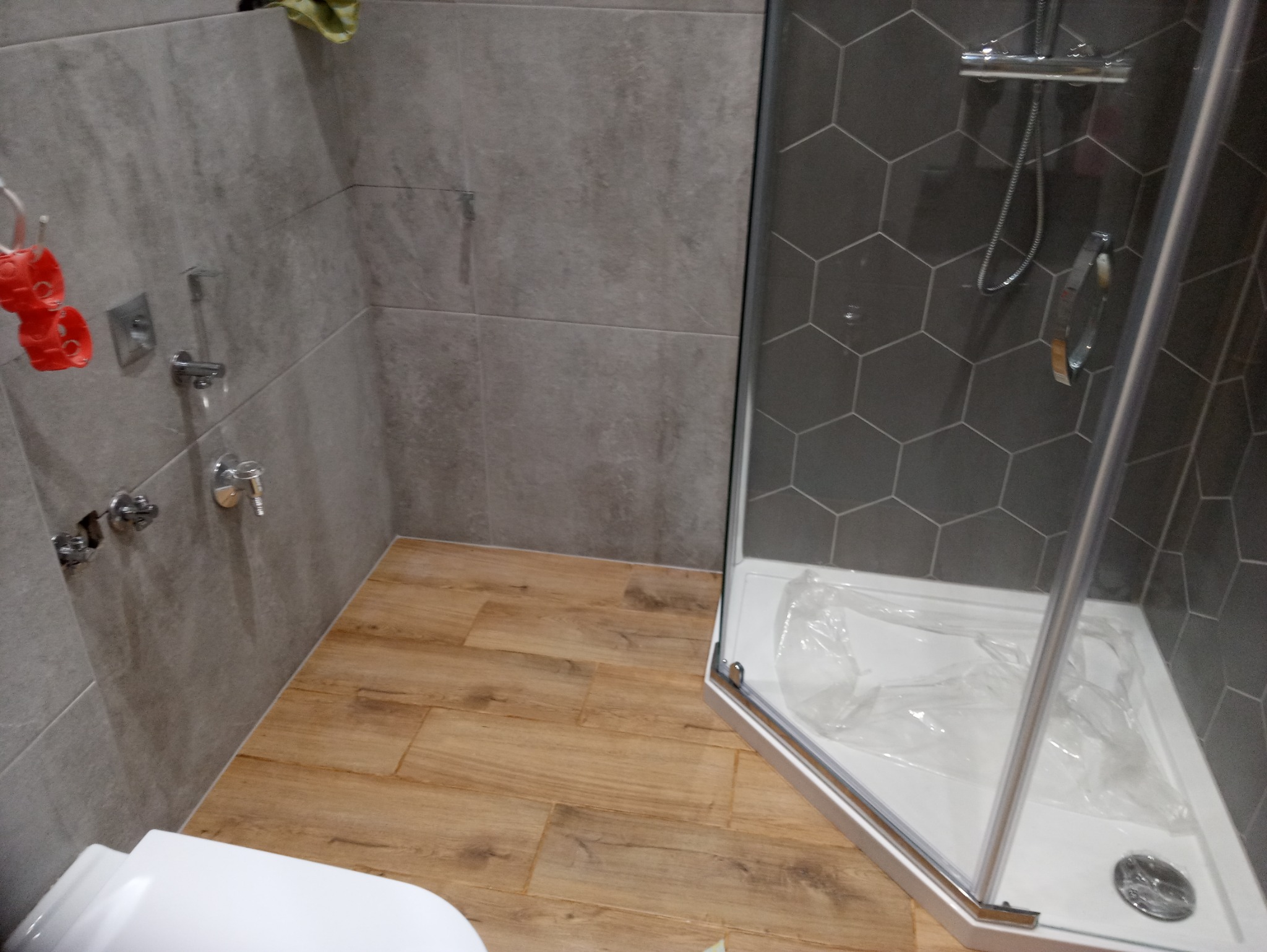 Wykończona łazienka z szarą, heksagonalną glazurą w kabinie prysznicowej, drewnopodobną podłogą i szarą ceramiką ścienną, widoczne chromowane baterie i akcesoria, kabina prysznicowa zabezpieczona...