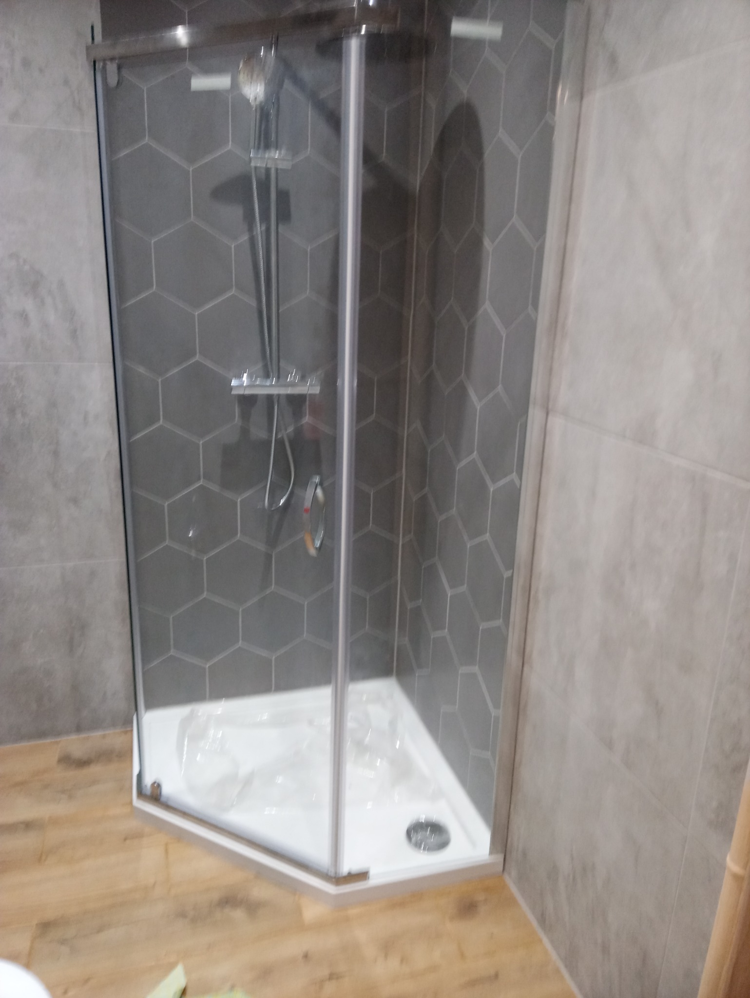 Narożna kabina prysznicowa z chromowaną armaturą na tle szarych płytek heksagonalnych i imitujących drewno płytek podłogowych.
