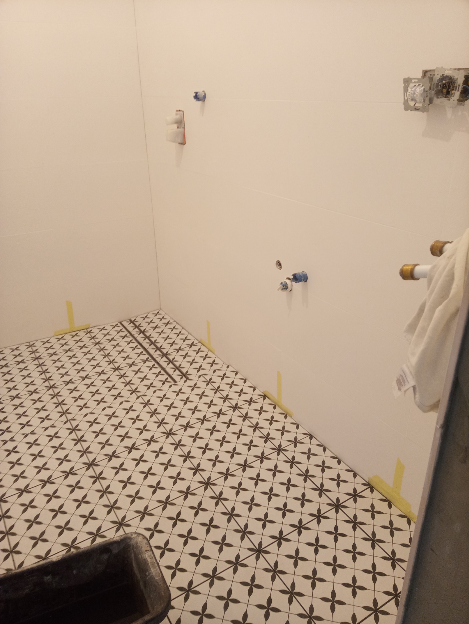 Łazienka w trakcie remontu: ułożone płytki podłogowe w biało-czarny wzór, widoczne instalacje wodne na ścianie i taśma malarska zabezpieczająca krawędzie.