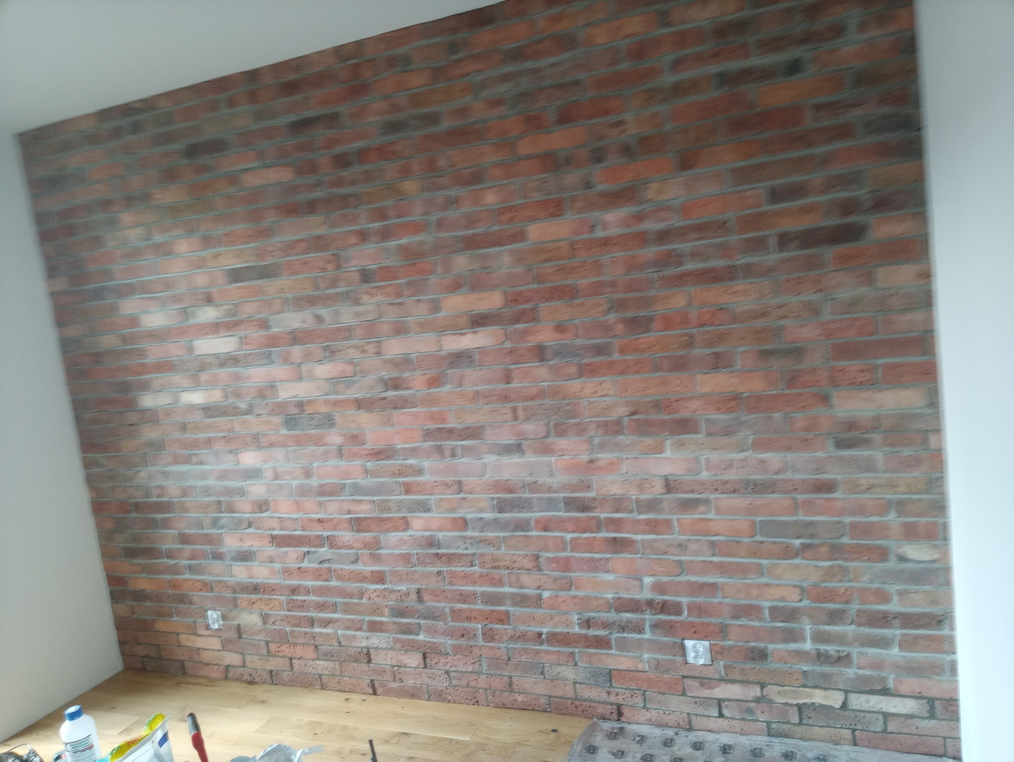 Ściana z cegieł klinkierowych w odcieniach czerwieni i brązu, z widocznymi fugami, fragment podłogi z jasnego drewna oraz białe ściany boczne, gniazdka elektryczne na ścianie.